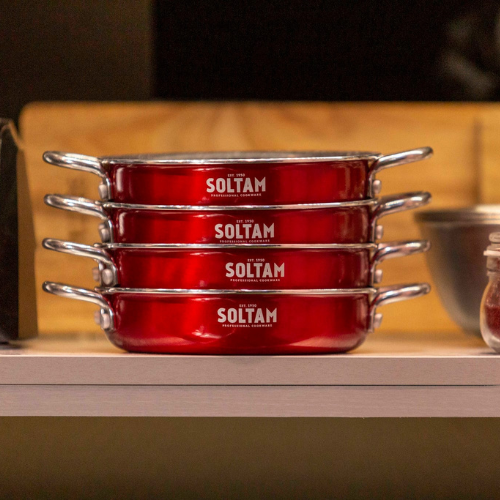 במרכז התמונה שלושה כלים צרים ועמוקים בצורת סוטאז׳ (מחבתות־סירים), בצבע אדום בוהק, מונחים זה על זה על מדף עץ. לכל כלי ידית מתכת מבריקה בצד ימין, ובמרכז דופן הכלים רשום בלבן שם המותג: “סולתם”. הרקע מאחורי המדף בצבע צהבהב מטושטש.